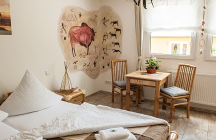 Ferienwohnung mit Doppelbett, einer gemalten Höhlenmalerei an der Wand und einem Esstisch mit zwei Stühlen am Fenster